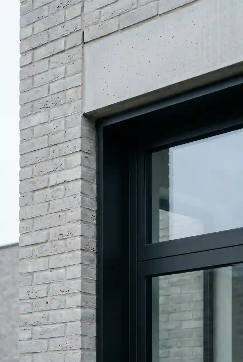 Junction of a pale grey brick wall, a deep-set dark aluminium window frame and a precast concrete lintel